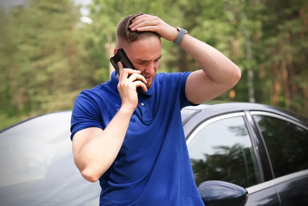 Stressed driver talking on the phone with an insurance adjuster beside his car after an accident