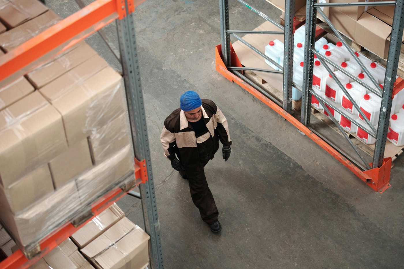A warehouse worker in a blue cap walks between shelves stacked with cardboard boxes and large bottles. The setting is industrial and organized.