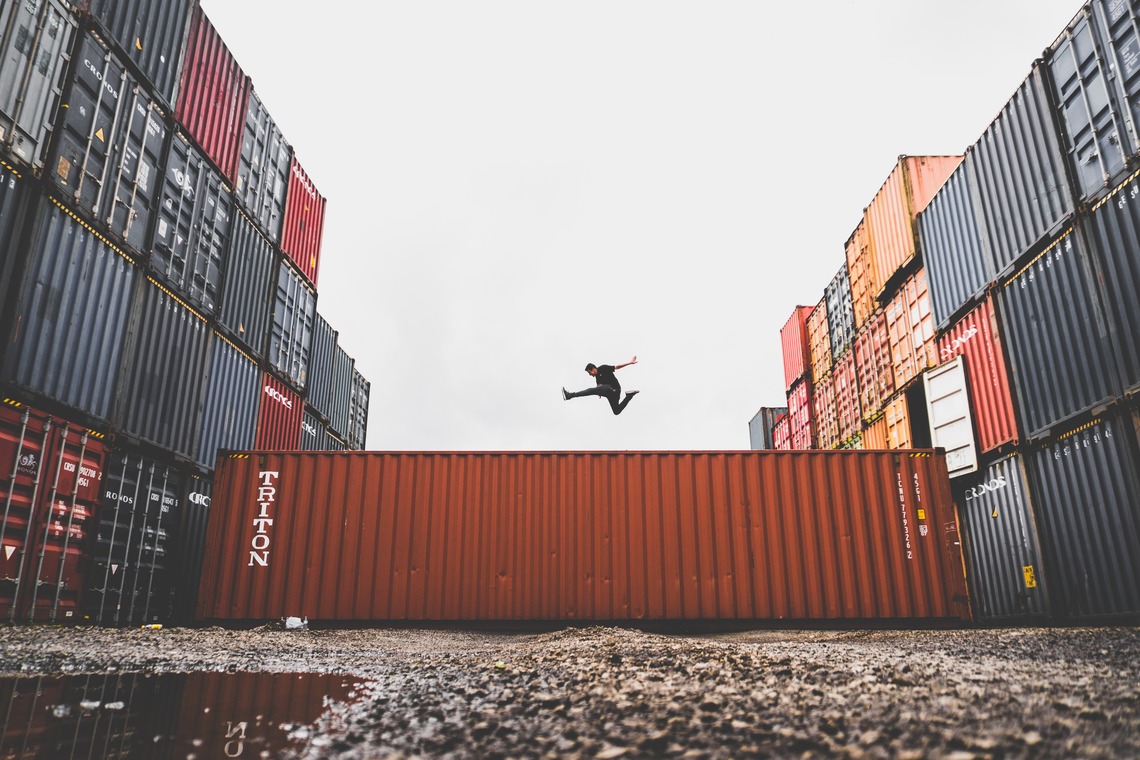 Person mid-air jumping between stacked shipping containers in an industrial yard.