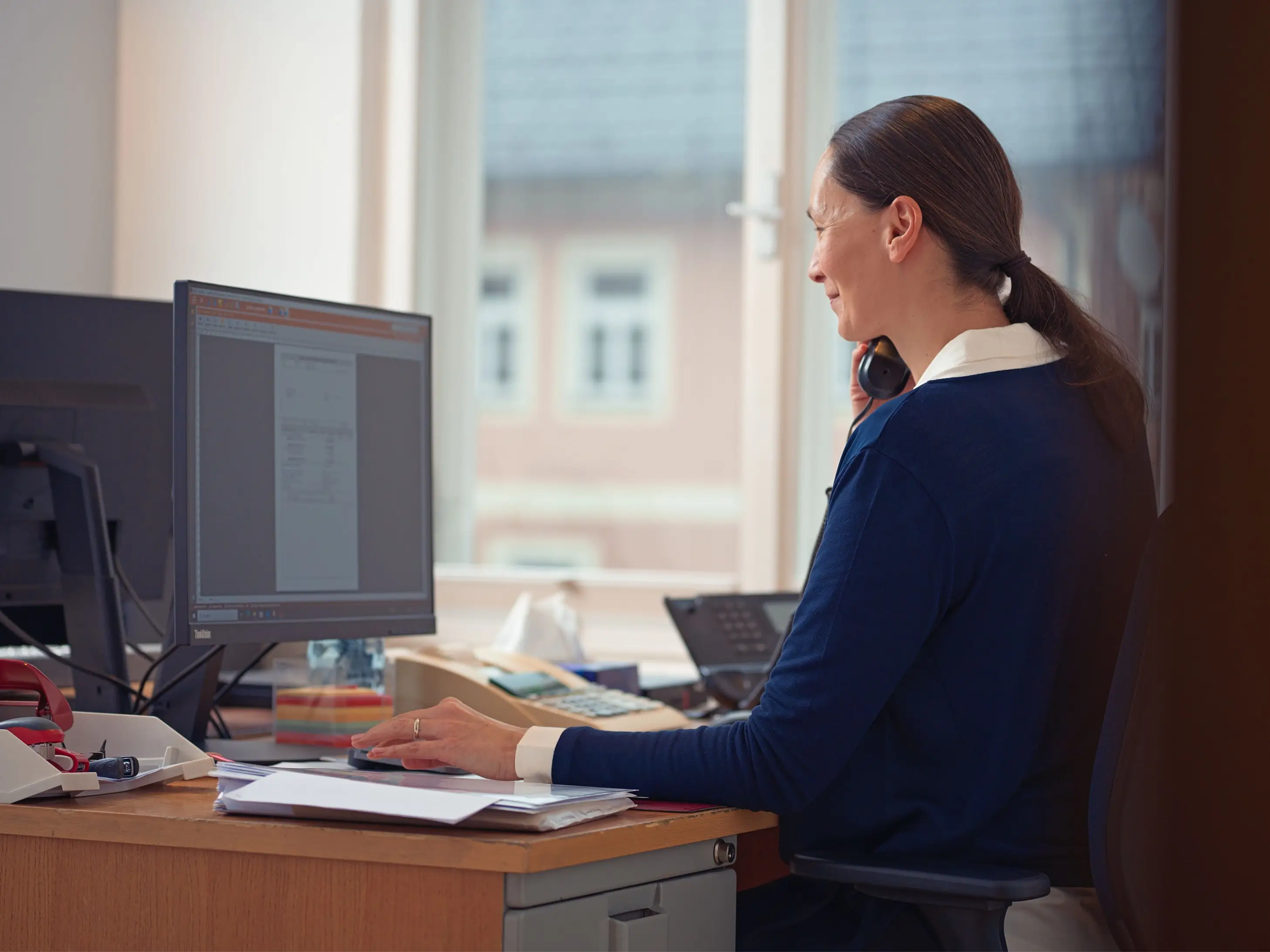 Frau sitzt an einem Schreibtisch, benutzt Computer und spricht am Telefon in einem hellen Büro mit Fenster.