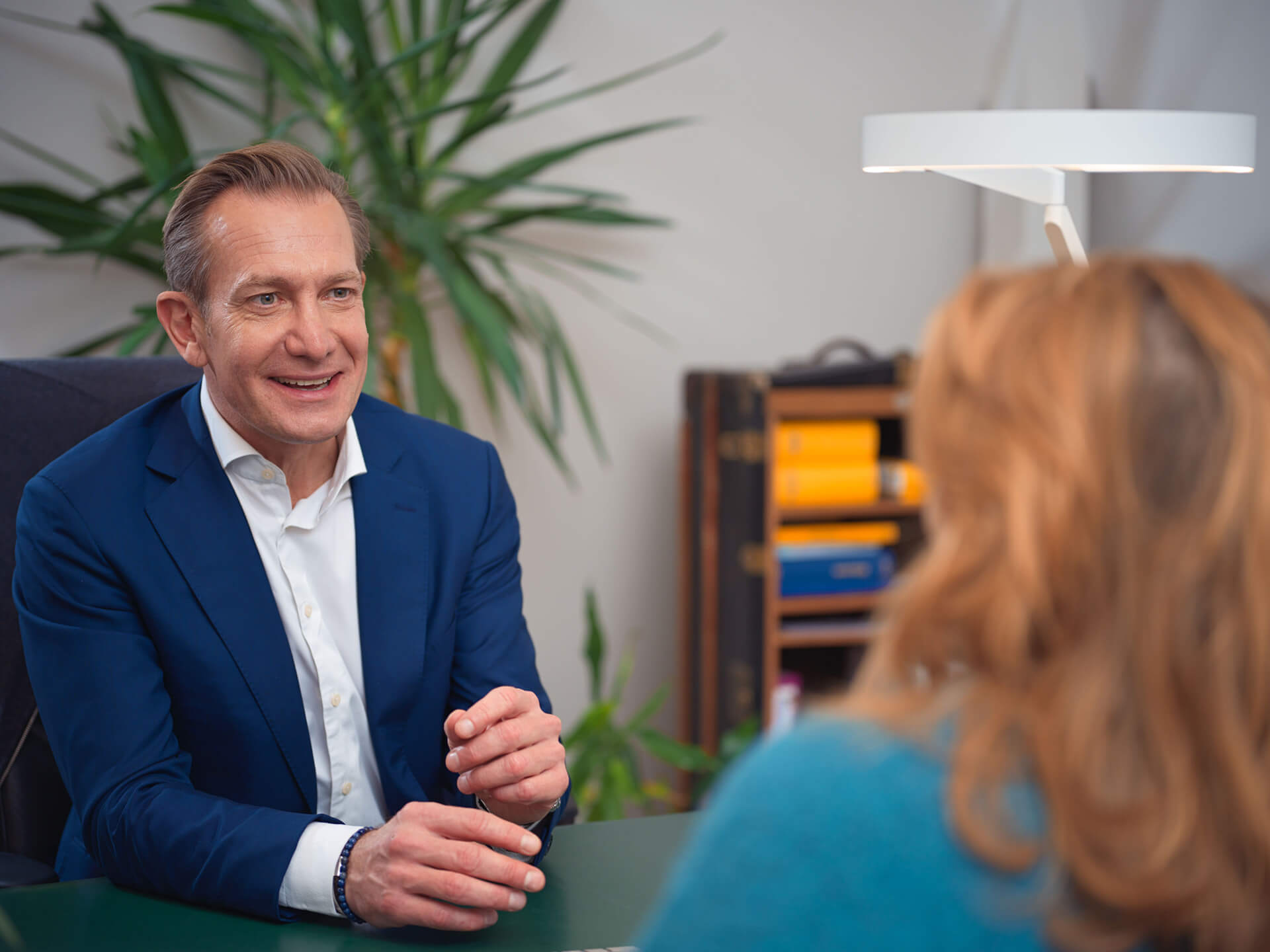 Mag. Dr. Christoph Mayrl – in blauem Anzug – spricht freundlich mit einer Frau mit rötlichen Haaren in einem Büro mit Pflanzen und Bücherregal im Hintergrund.