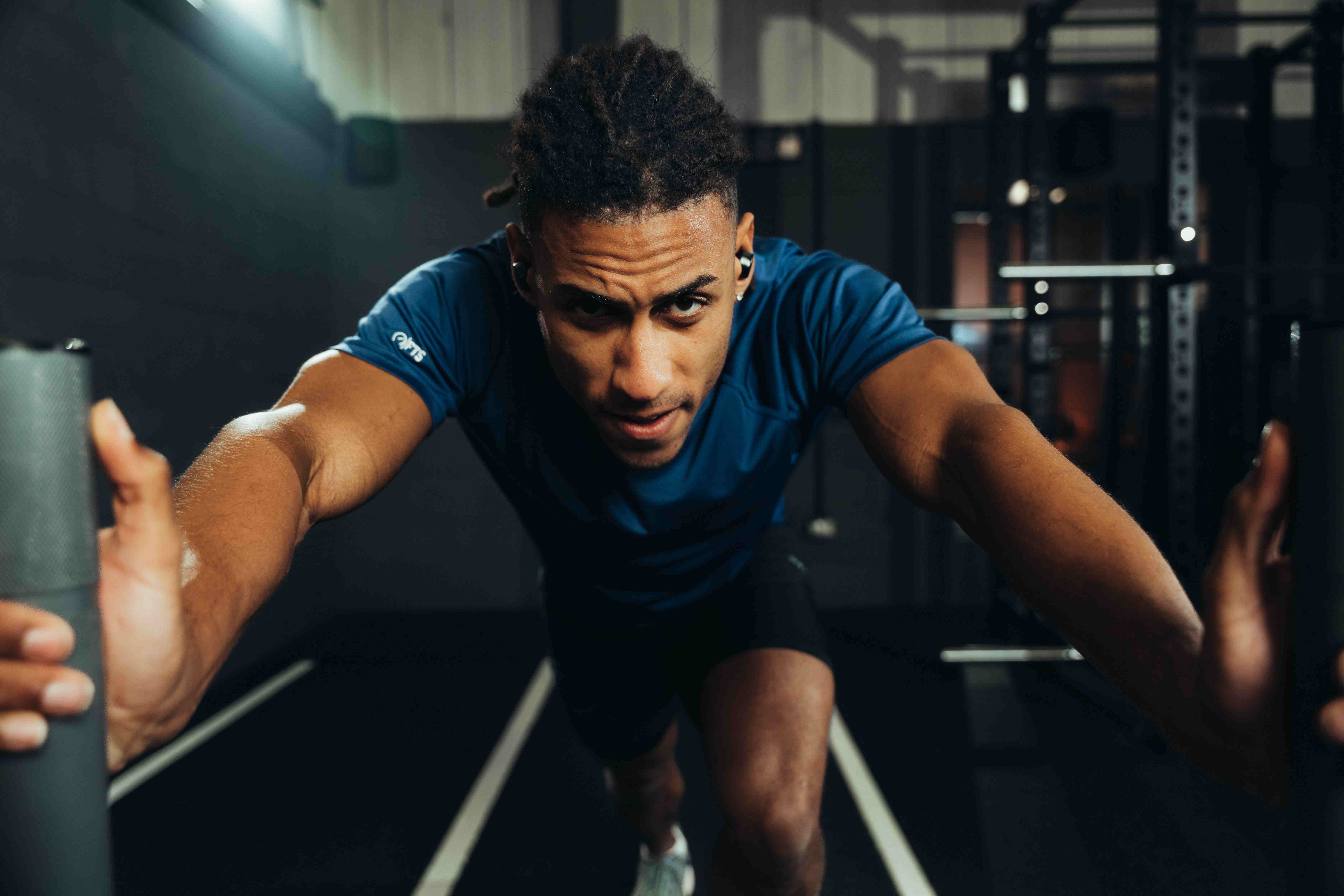 A man in a blue athletic t-shirt in a sprinting start position on an indoor gym track, looking intensely at the camera.