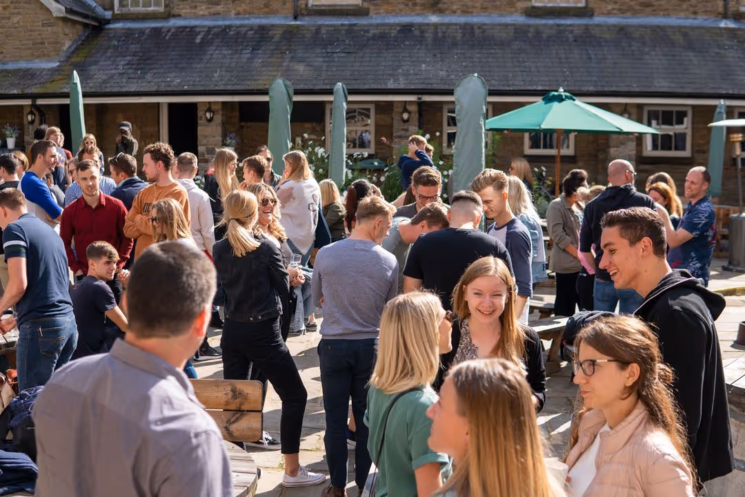 Group of young adults socializing outdoors near picnic tables with umbrellas and closed parasols.