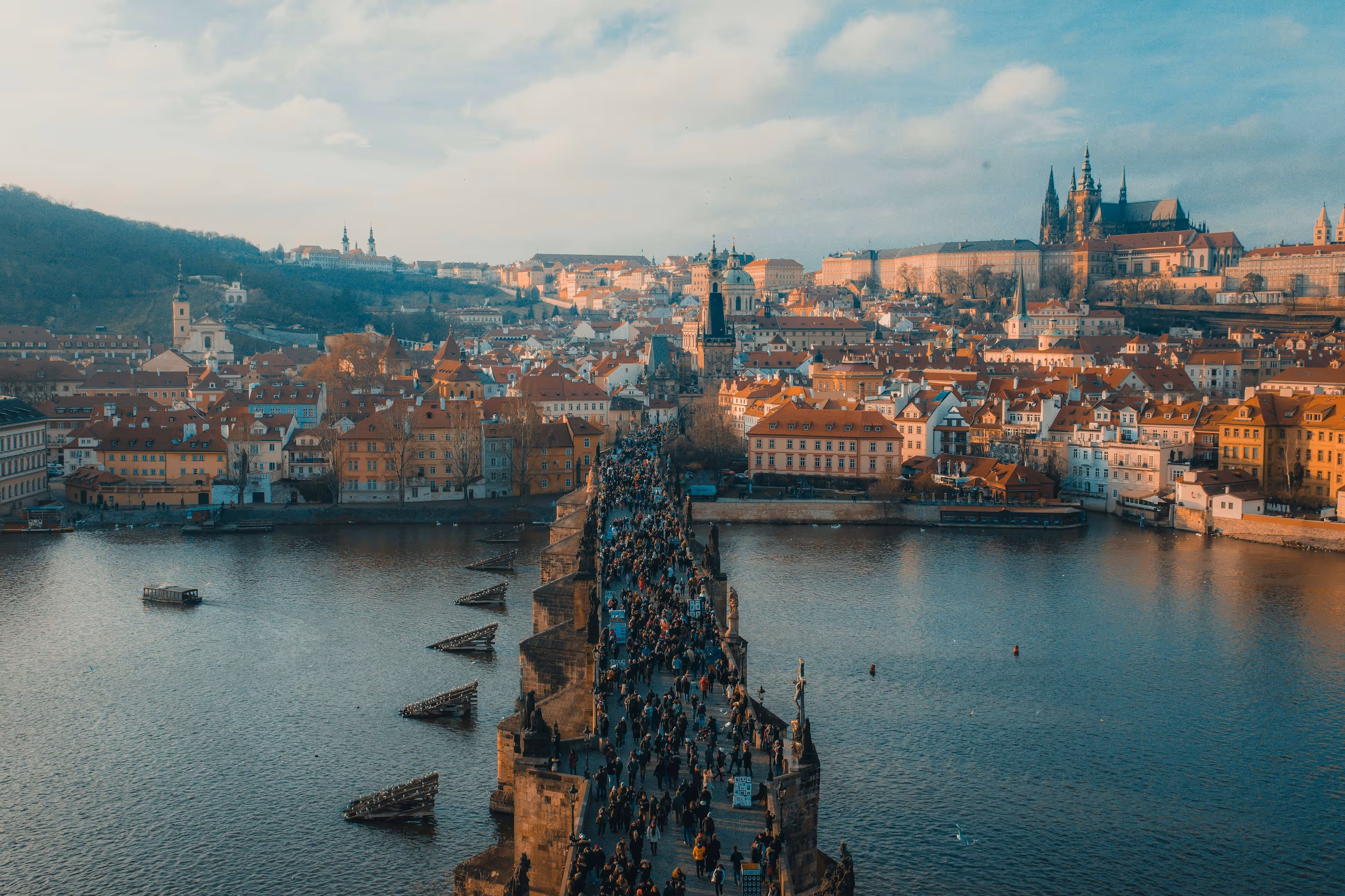 A crowded Charles Bridge over the Vltava River with historic buildings and Prague Castle in the background during sunset.