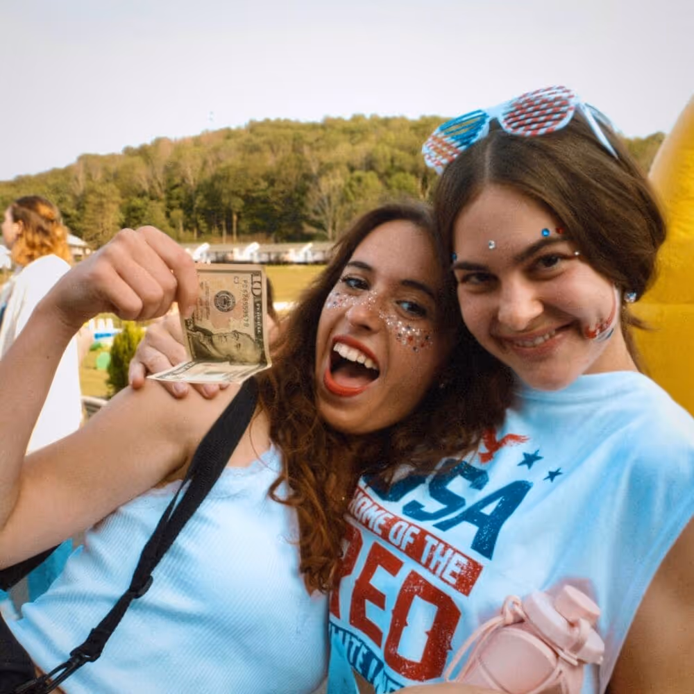 Two young women smiling outdoors, one holding a ten-dollar bill and the other wearing patriotic sunglasses and holding a pink water bottle.