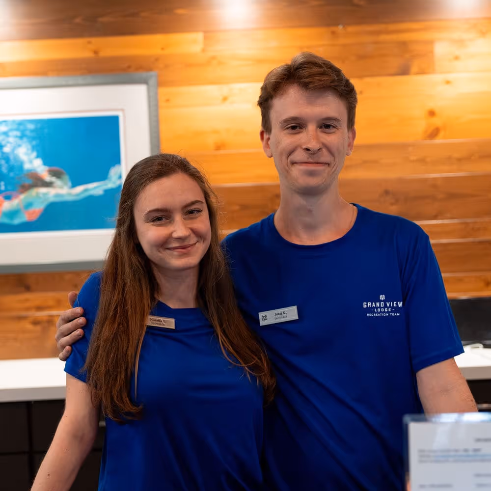 Two smiling employees wearing blue Grand View Lodge shirts stand side by side in a wood-paneled reception area.