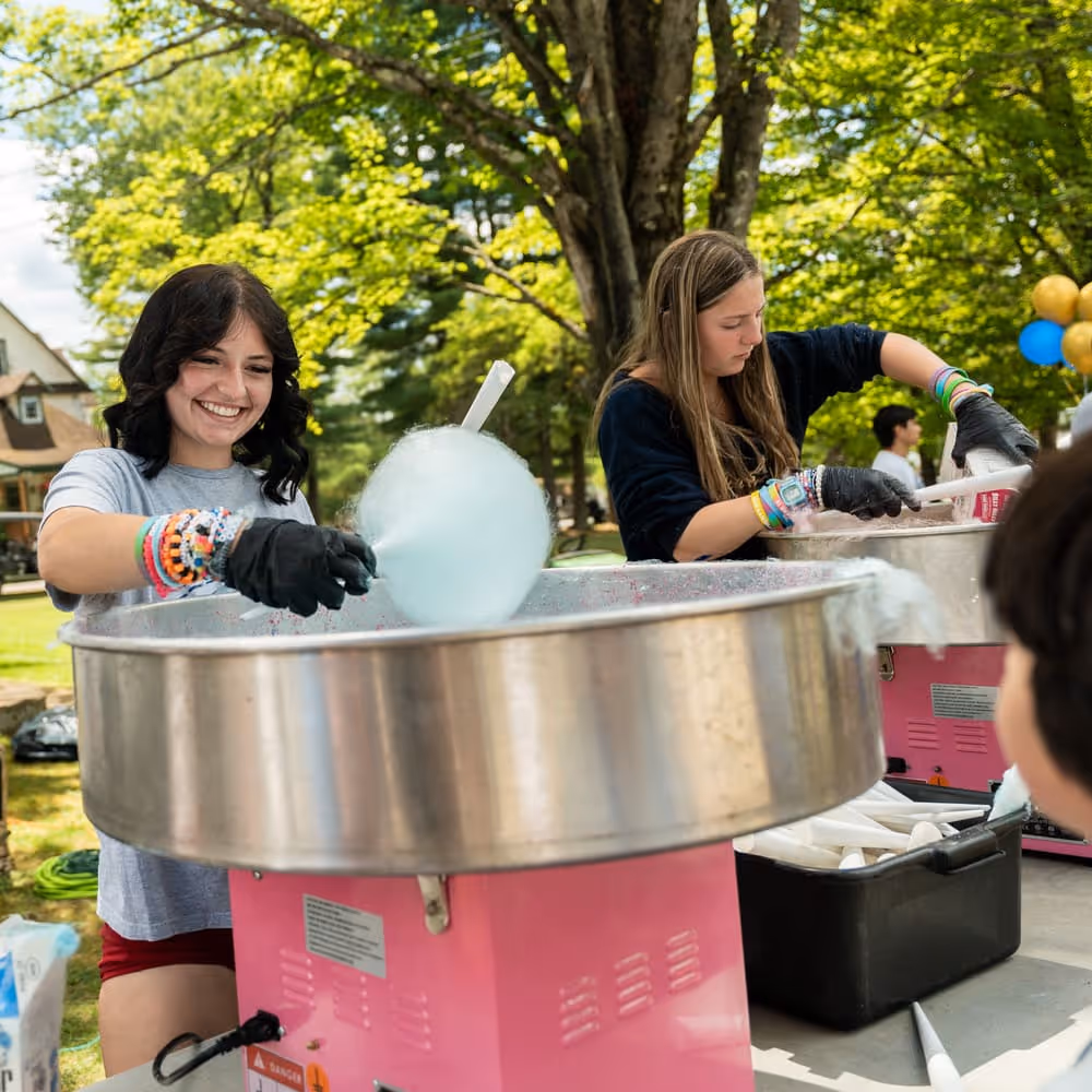Two young women making cotton candy outdoors on sunny day, one smiling while spinning blue cotton candy on a stick.