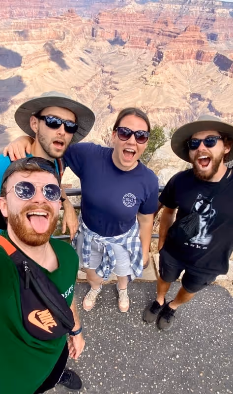 Four friends wearing sunglasses and hats smiling and posing for a selfie at the Grand Canyon viewpoint.