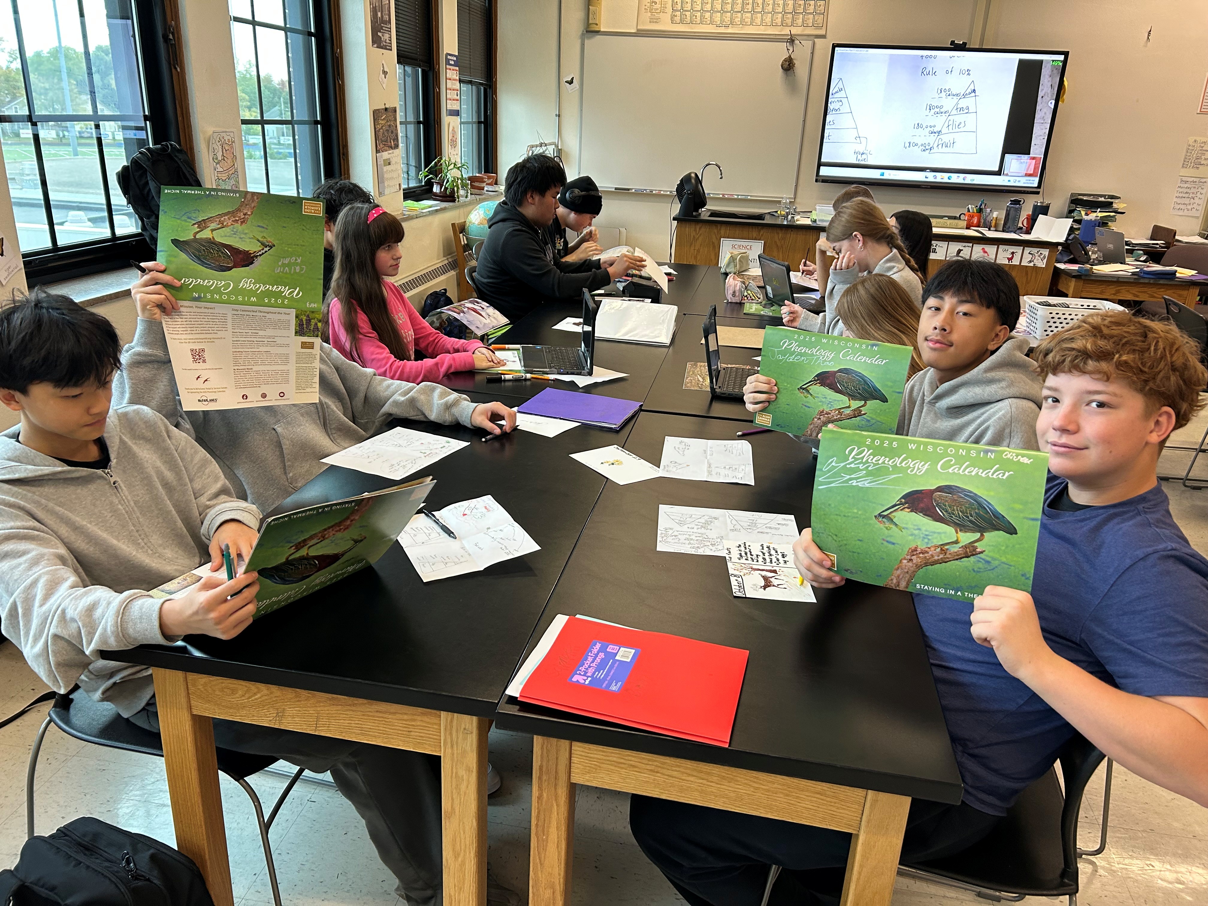 Students sitting around a lab table in a science classroom hold up wildlife calendars.