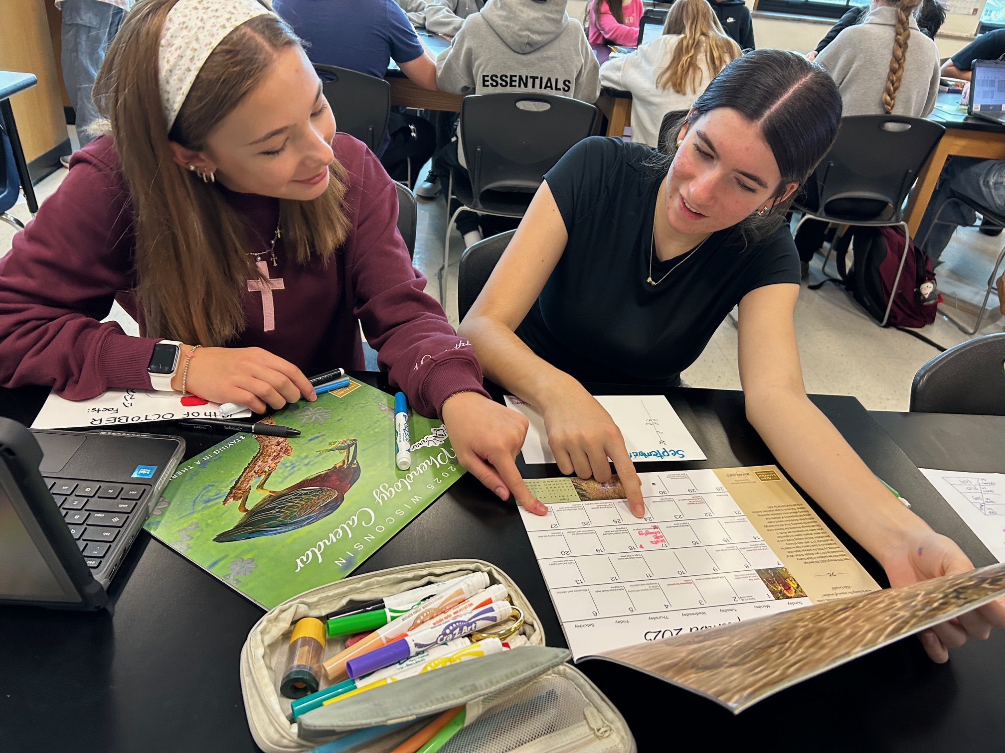 Two girls sitting at a lab table point to dates on a calendar grid.