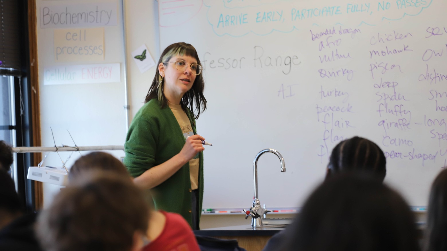 A woman with dark hair and glasses stands at the front of a classroom in front of a whiteboard with a list of words written on it.