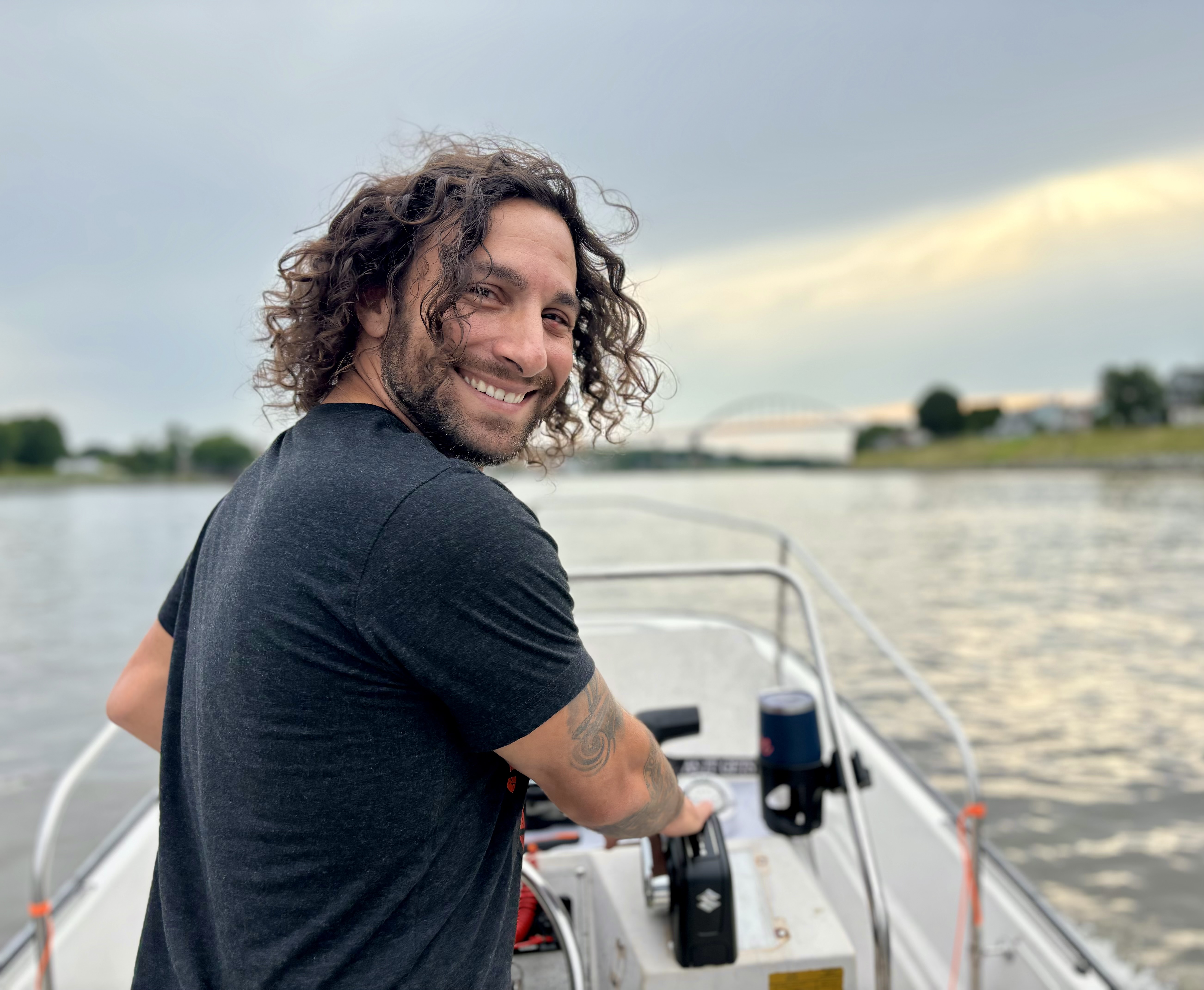A bearded man with curly hair looks back at the camera from his position at the stern of a small motorboat.