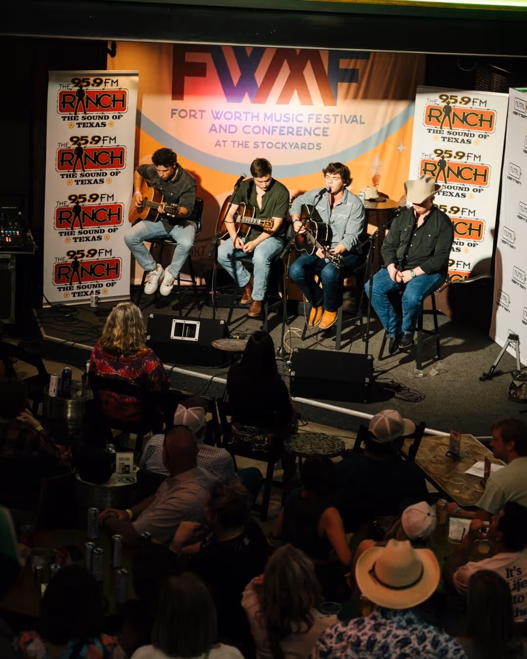 Four musicians seated on stools performing with acoustic guitars on stage at the Fort Worth Music Festival and Conference, with an audience watching.