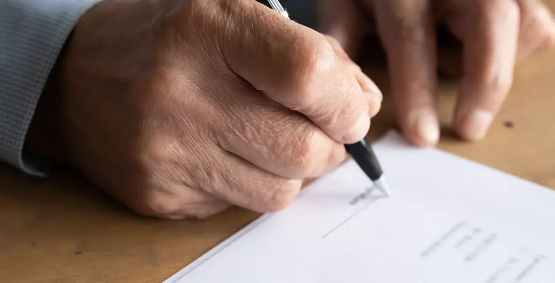 Close-up of an elderly person's hand signing a document with a black pen on a wooden table.