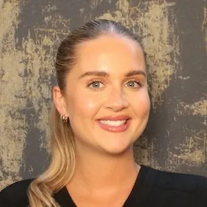 Smiling woman with light skin, blue eyes, and blonde hair pulled back, wearing a black top against a textured gray background.