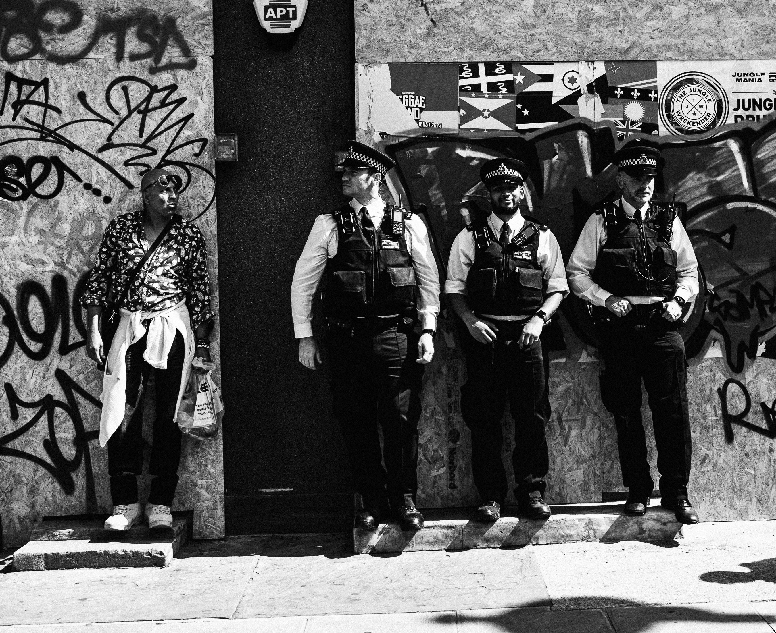A festivalgoer stands apart from a line of police officers against a graffiti-covered wall at Notting Hill Carnival.