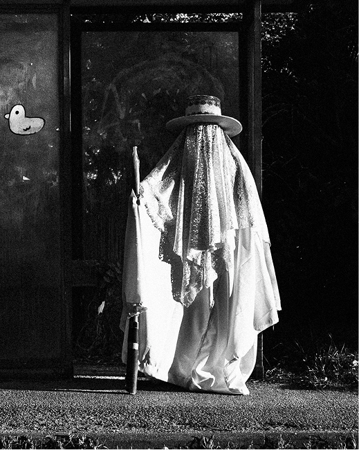 A white-robed Eyo figure stands beside a weathered bus shelter on a London street.