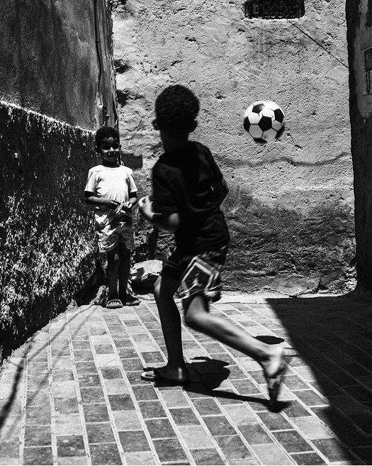 Two children play football in a narrow sunlit passage in Morocco, with the ball suspended between them.