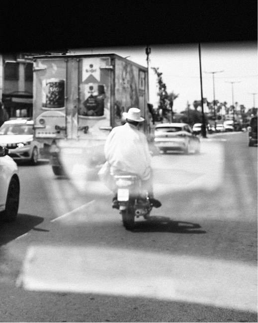 Cars, passengers and a motorcyclist overlap in reflections seen through a vehicle window in Morocco.