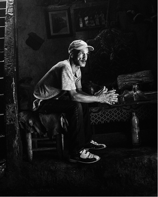 A seated man wearing a cap rests his hands together inside a dark Moroccan interior illuminated by natural light.
