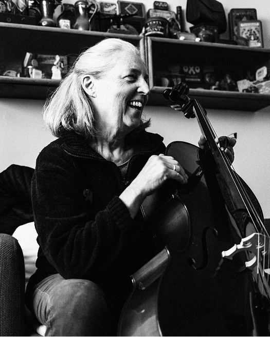 A cellist laughs while holding her instrument inside a room filled with personal objects.