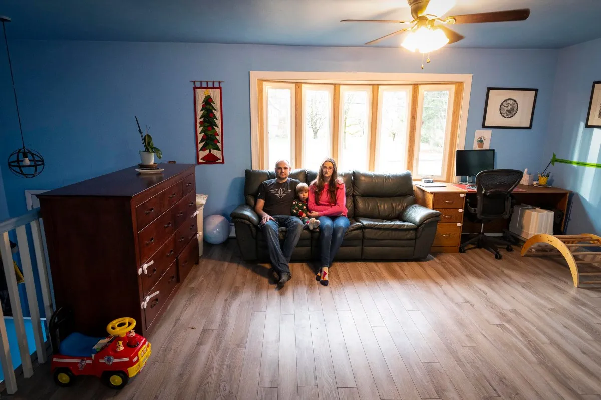 A couple with a child sits on a couch in a living room. The room has blue walls, a ceiling fan, a desk, and a toy car on the floor.