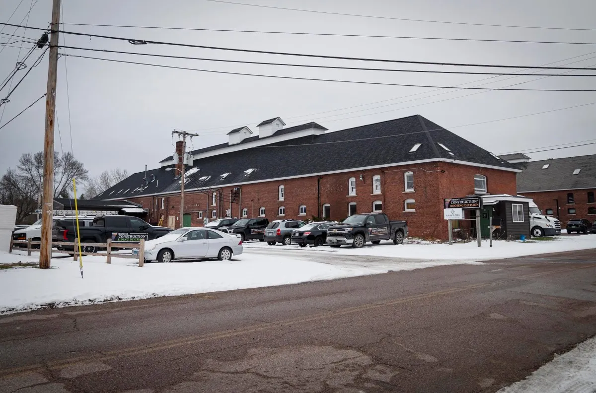 Large brick building with a dark roof, surrounded by parked cars. Snow covers the ground and rooftops. Power lines are visible overhead. A sign near the building indicates a business entrance.