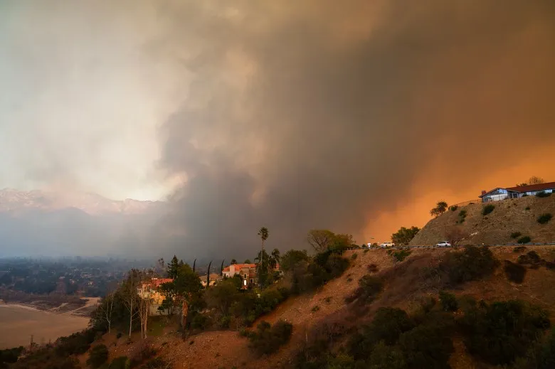 A hillside neighborhood with scattered houses and trees is illuminated by an orange glow, as thick smoke fills the sky. Mountains are visible in the background under a hazy, pale sky. A road with a few parked cars runs along the edge of the hill.