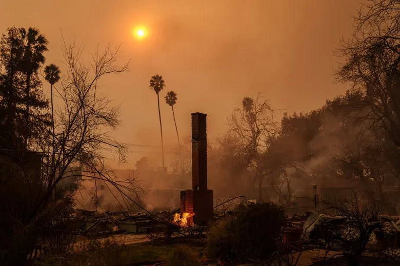 A fireplace standing over the rubble and debris from a home that the Eaton Fire in Altadena burned down. Smoke from the nearby fire is blocking the sun in the background with palm trees and debris from a destroyed neighborhood.