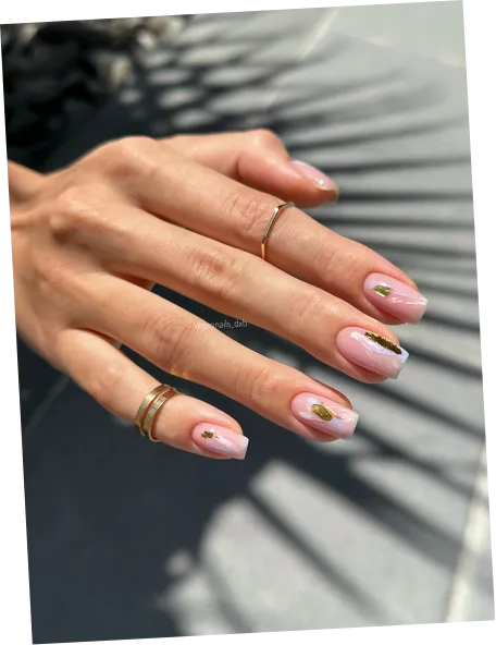 Hand with pink nails featuring gold leaf accents and rings on fingers over shadowed background.
