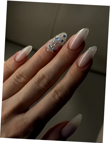 Hand with long almond-shaped nails in soft pink ombre, one nail decorated with multicolored rhinestones.