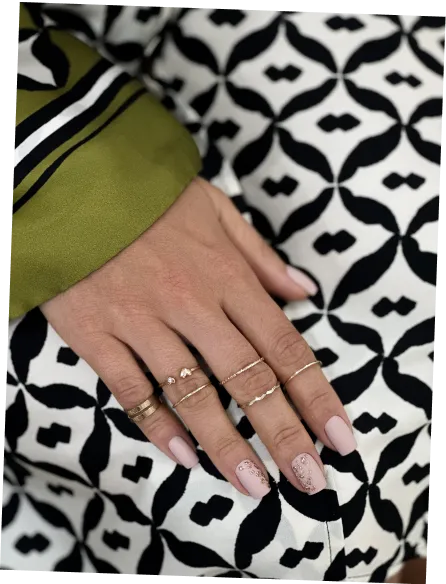 Hand with light pink nail polish and delicate gold rings resting on black and white patterned fabric.