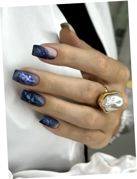 Hand with purple and black marble-effect nails and a large gold ring with a white pearl.