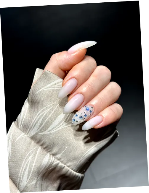 Hand with long, almond-shaped nails in light pink and one nail adorned with blue gemstones.
