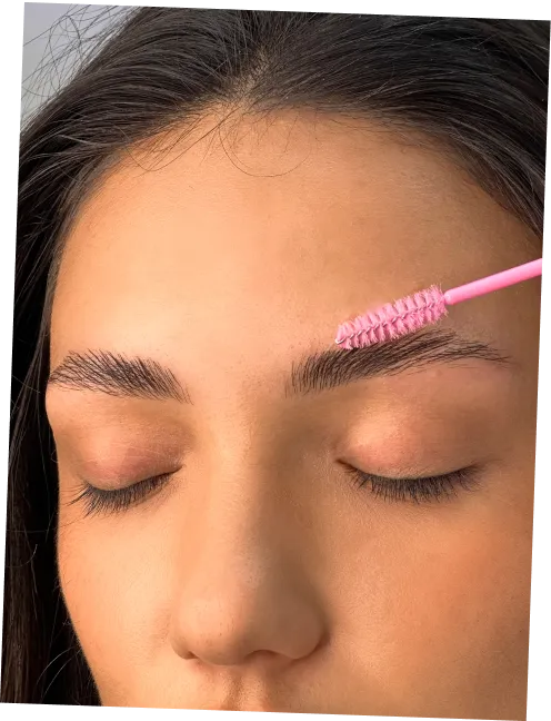 Close-up of a person with closed eyes grooming an eyebrow with a pink spoolie brush.