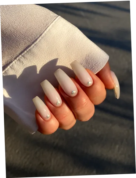 Hand with long matte nude coffin-shaped nails, each nail adorned with a small shiny gem near the cuticle.
