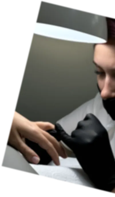 Person wearing black gloves applies nail treatment to another person's hand under a lamp.
