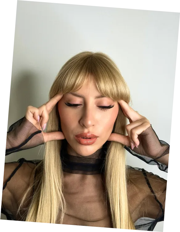Woman with long blonde hair in sheer black top with eyes closed, fingers touching temples, posing against white wall.