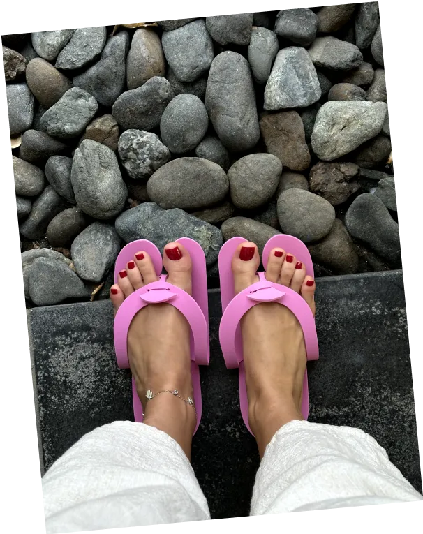 Feet with red nail polish wearing pink sandals standing on dark stone floor with pebble stones background.