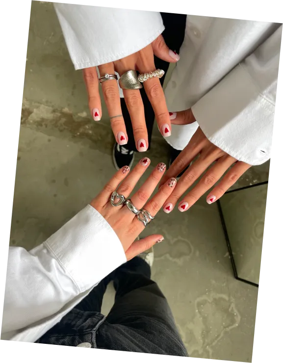 Two hands with white nail polish and red hearts, one with leopard print tips, both wearing silver rings.
