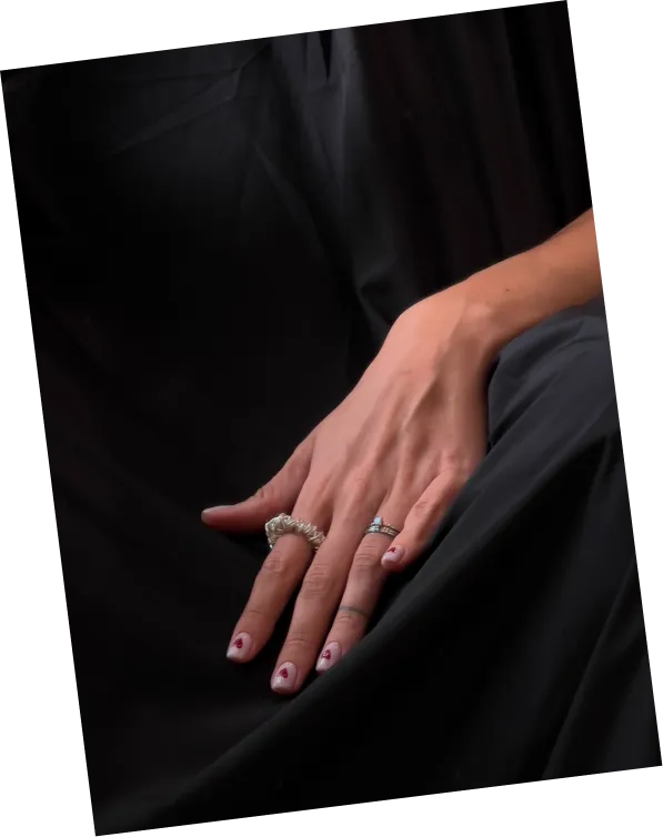 A hand with two rings and heart-decorated nails resting on a black fabric background.