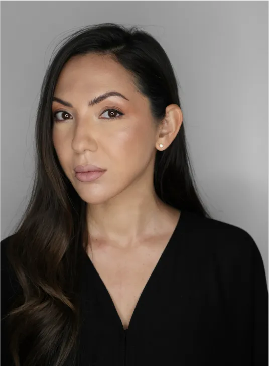 Portrait of a woman with long dark hair and pearl earrings wearing a black top against a gray background.