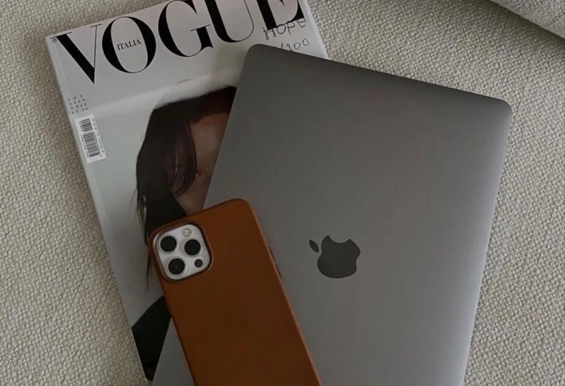 Closed Apple MacBook and iPhone with brown case resting on Vogue Italia magazine on a textured fabric surface.