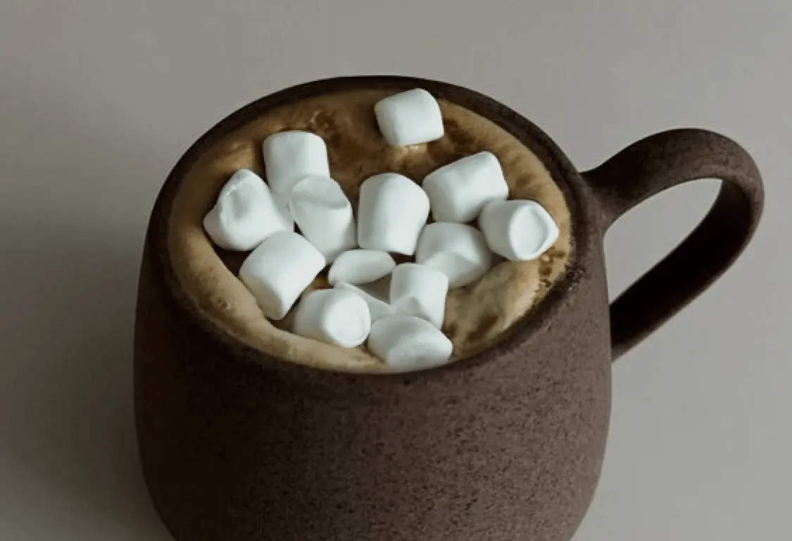 Brown mug filled with hot chocolate and topped with small white marshmallows on a gray surface.