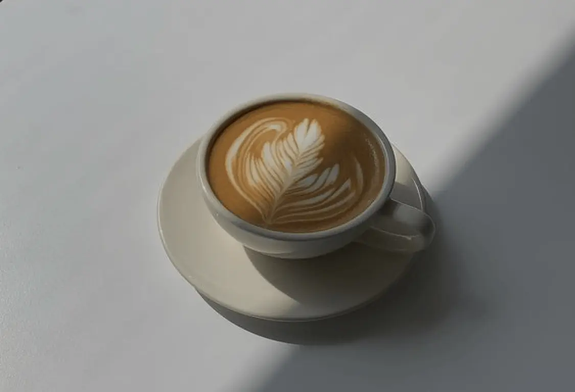 Cup of latte coffee with leaf-shaped foam art on a saucer casting a shadow on a light surface.