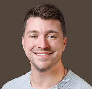 Portrait of a smiling man with short brown hair wearing a light gray crewneck sweater, set against a solid dark background.