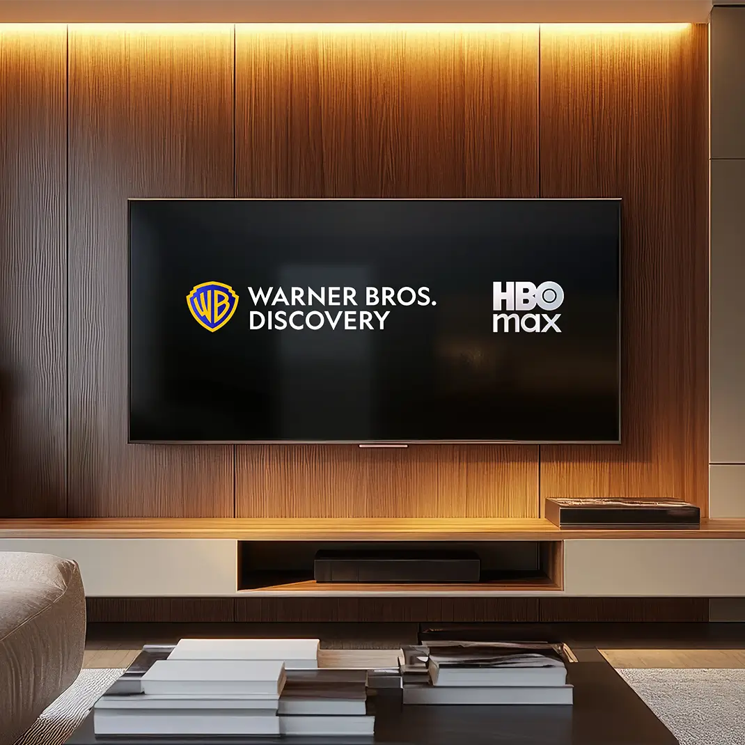 The logos for Warner Bros. Discovery and HBO Max sit side-by-side, showcased on a sleek flatscreen TV in a living room. The warm glow of track lights above and below a wooden alcove frames the TV, which is set in a sleek, modern living room above built-in media storage. In the foreground are the edge of a cream-colored couch and a coffee table with stacks of books on top.