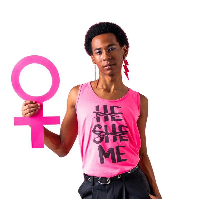 A person with curly hair and pink lightning bolt earrings holds a large pink Venus symbol (♀) in their left hand. They wear a bright pink tank top with the words "HE" at the top, "SHE" in the middle struck through with a diagonal line, and "ME" at the bottom, paired with black pants and a belt, standing against a black background. This image illustrates the personal and evolving nature of gender identity.