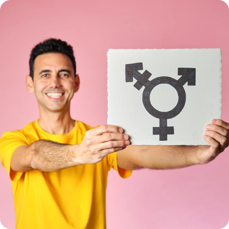 A smiling person with short dark hair, wearing a bright yellow t-shirt, holds up a white sign featuring the transgender symbol—a combination of male (♂) and female (♀) symbols with an additional arrow—against a soft pink background. This image represents the diversity and spectrum of gender identities beyond traditional binaries.
