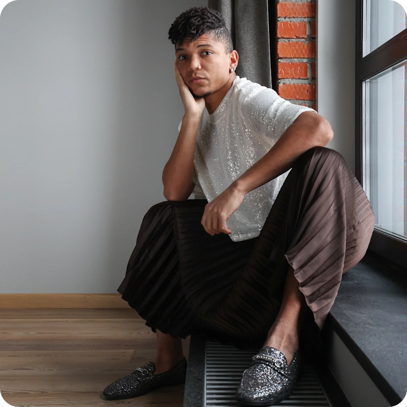 A thoughtful person with short curly hair and an earring sits on a windowsill, chin resting on their hand, wearing a sparkly white short-sleeve shirt, brown pleated pants, and glittery silver loafers, in an indoor setting with a brick wall and wooden floor. This image conveys the emotional discomfort and introspection often associated with gender dysphoria.